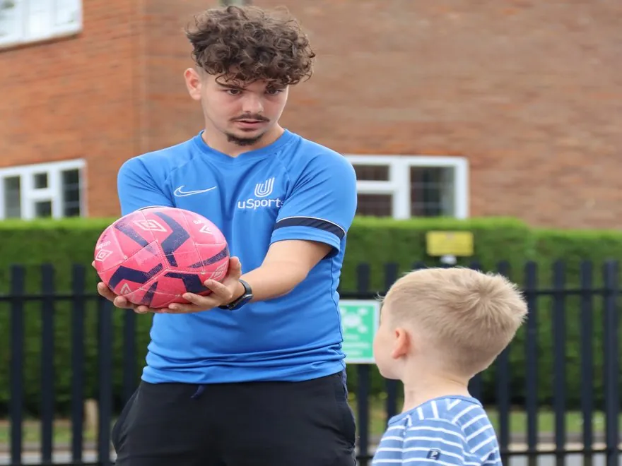 A uSports coach handing a football to a young child at a school session